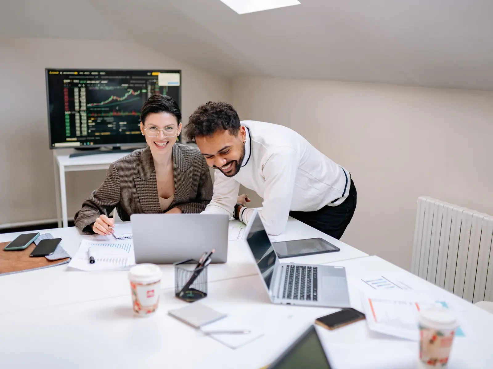 A man leaning over a colleague's desk as they both smile at her laptop
